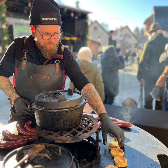Ein bärtiger Mann in einer Schürze kocht Würste und Pfannkuchen auf einem Grill im Freien. Er hat einen Topf und einen Holzlöffel in der Nähe. Im Hintergrund stehen und gehen Leute.