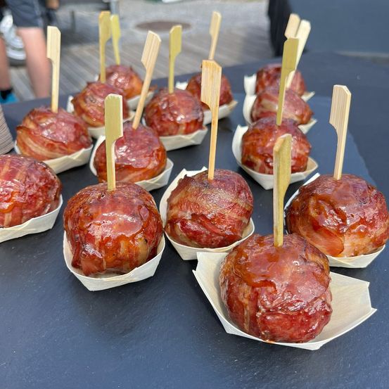Ein Tisch mit mehreren in Speck gewickelten Fleischbällchen auf Papiertellern mit Holzstäbchen. Die Fleischbällchen sind glasiert und sehen saftig aus.