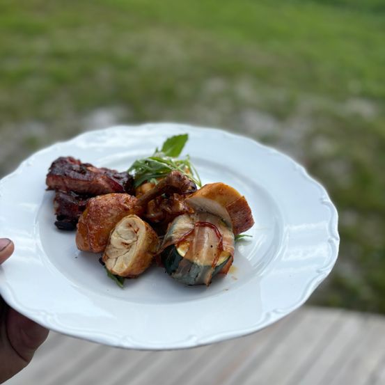 Ein weißer Teller hält ein Gericht mit gegrilltem Fleisch, Gemüse und Kräutern, vor einem verschwommenen Grashintergrund.
