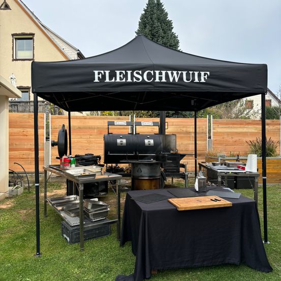 Ein Grill-Setup im Freien mit einem schwarzen Vordach, beschriftet mit 'Fleischwuif'. Ein Grill und Kochutensilien befinden sich unter dem Vordach. Ein Holzzaun und Bäume sind im Hintergrund.