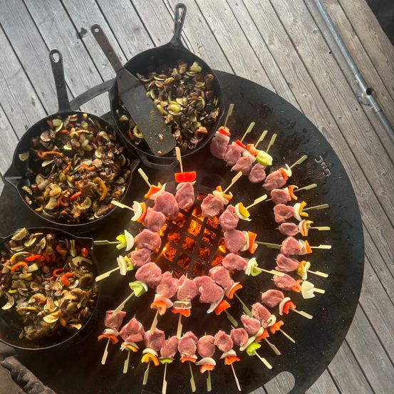 Ein schwarzer Grill mit mehreren Spießen mit Fleisch und Gemüse darauf. Schüsseln mit Essen sind um den Grill herum, und eine wird darauf gekocht.