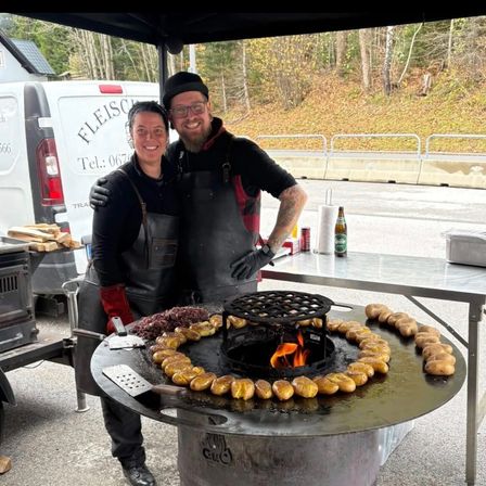 Ein Paar steht hinter einem Grill mit Feuerstelle und kocht Kartoffeln im Freien. Ein weißer Van und ein Tisch mit einer Flasche sind in der Nähe.
