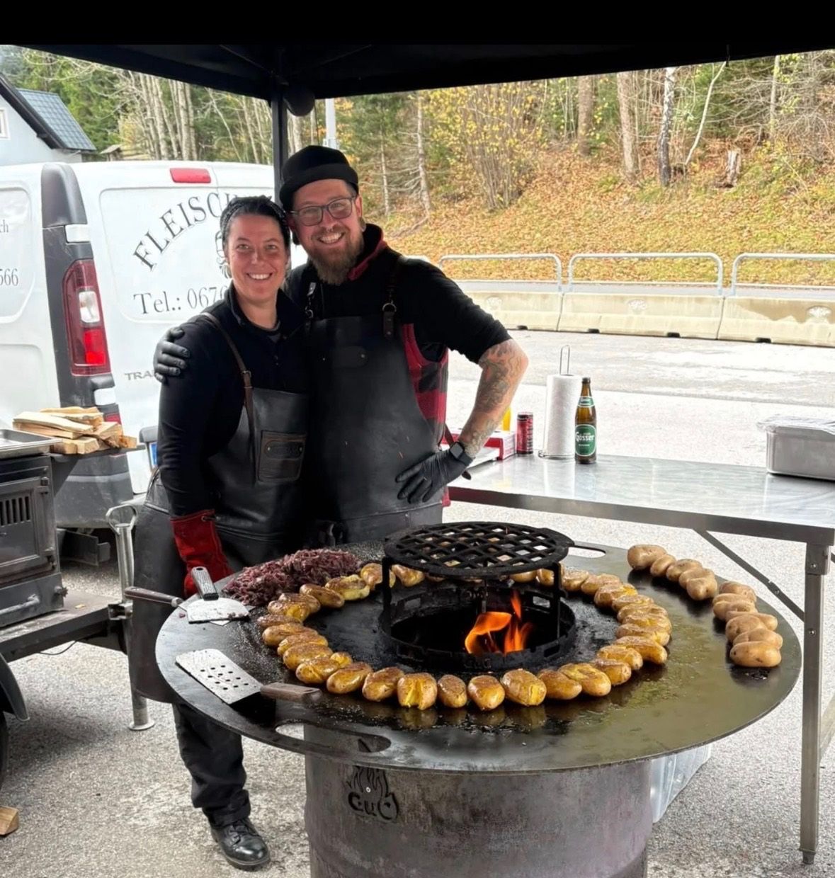 Ein Paar steht hinter einem Grill mit Feuerstelle und kocht Kartoffeln im Freien. Ein weißer Van und ein Tisch mit einer Flasche sind in der Nähe.