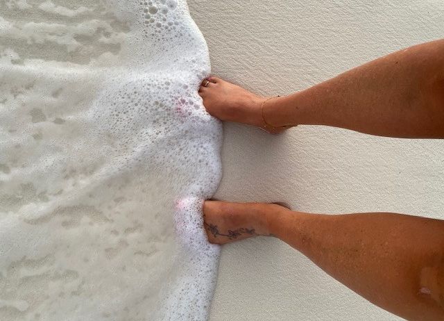 Eine Person steht barfuß auf einem sandigen Strand, mit einem Fuß im schaumigen Wasser. Sie hat ein Tattoo am Knöchel.