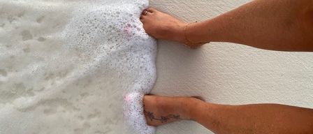 Eine Person steht barfuß auf einem sandigen Strand, mit einem Fuß im schaumigen Wasser. Sie hat ein Tattoo am Knöchel.