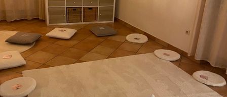 Ein ruhiger Meditationsraum mit einem großen weißen Teppich, runden Kissen und dekorativen Objekten. Ein Schrank mit Schubladen und Regalen, eine Uhr an der Wand und ein Fenster mit Vorhängen.