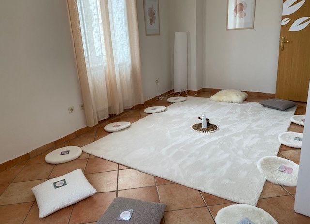 Ein ruhiger Raum mit einem weißen Teppich, mehreren weißen Kissen und einer Holztür. Der Raum hat ein minimalistisches Design mit einer weißen Wand, einem Fenster mit Vorhängen und einer hohen weißen Lampe. Es gibt zwei gerahmte Bilder an der Wand.