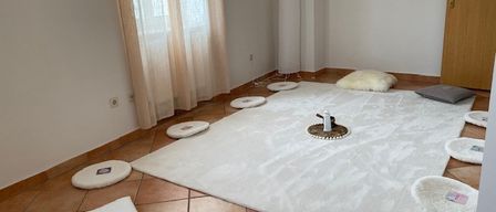 A serene room with a white rug, several white pillows, and a wooden door. The room has a minimalist design with a white wall, a window with curtains, and a tall white lamp. There are two framed pictures on the wall.