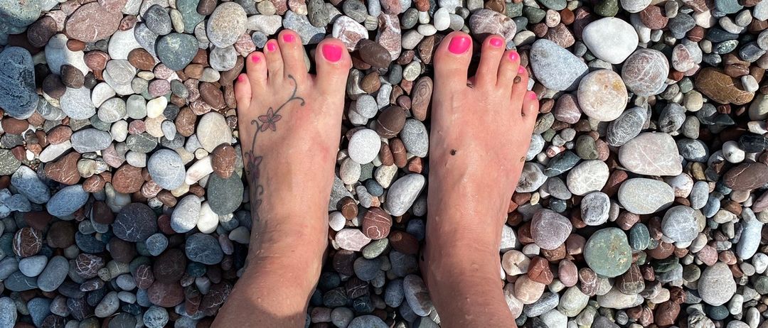 Eine Person mit rosa Zehennägeln steht barfuß auf einem Kieselstrand mit einem Tattoo auf dem linken Fuß.