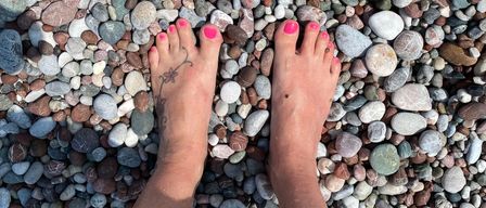 Eine Person mit rosa Zehennägeln steht barfuß auf einem Kieselstrand mit einem Tattoo auf dem linken Fuß.