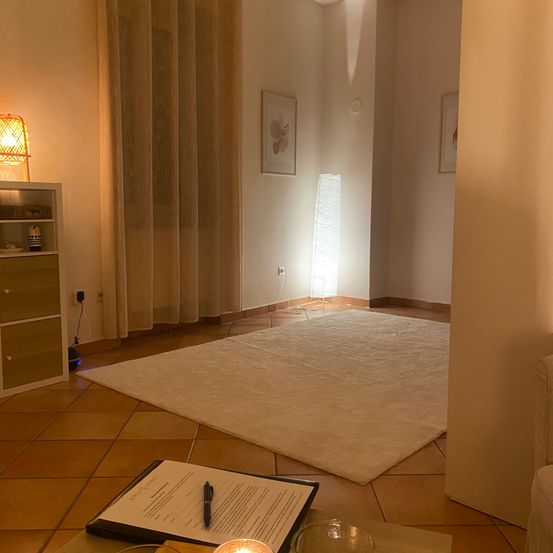 Ein gut beleuchteter Raum mit einem weißen Teppich, einem kleinen Tisch und einer leuchtenden Lampe. Es gibt einen weißen Schrank mit Regalen und einer Stehlampe. Ein Bilderrahmen ist an der Wand montiert.