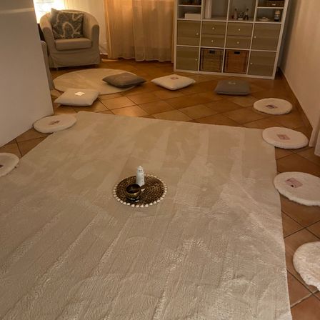Ein Zimmer mit einer beigefarbenen Couch, einem weißen Teppich und einem gefliesten Boden. Es gibt einen weißen runden Teppich in der Mitte mit einer weißen Kerze auf einem Teller.