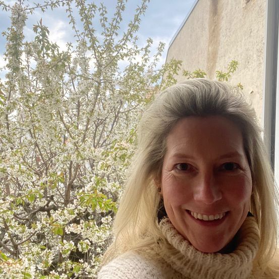 Eine lächelnde Frau mit grauem Haar steht vor einem Baum mit weißen Blüten. Sie trägt ein weißes Rollkragenshirt und scheint durch ein Fenster nach draußen zu schauen.