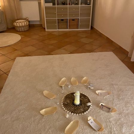 Ein weißer Teppich auf einem gefliesten Boden hat eine Kerze in der Mitte mit Blütenblättern darum. Kleine Steine sind verstreut. Ein Schrank mit Schubladen steht hinter dem Teppich.