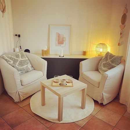 Ein gemütlicher Wohnbereich mit zwei weißen Stühlen, einem Holztisch und einem weißen Teppich. Ein Bilderrahmen, eine Lampe und eine Kerze befinden sich auf dem Regal. Taschentücher und eine Steckdose sind in der Nähe.