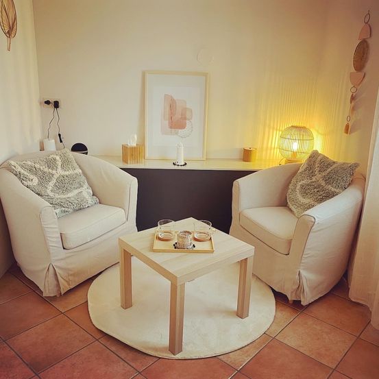 Ein gemütlicher Wohnbereich mit zwei weißen Stühlen, einem Holztisch und einem weißen Teppich. Ein Bilderrahmen, eine Lampe und eine Kerze befinden sich auf dem Regal. Taschentücher und eine Steckdose sind in der Nähe.