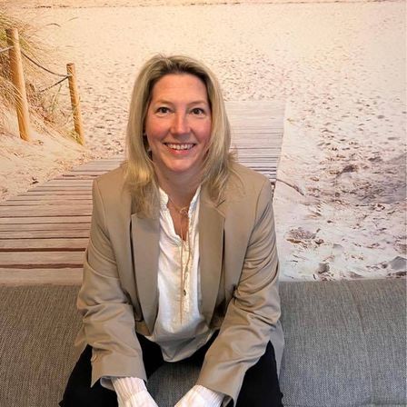 Eine lächelnde Frau in einem hellbeigen Anzug sitzt vor einem Strandhintergrund auf einem Sofa.