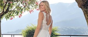 Eine Frau in einem weißen Hochzeitskleid steht draußen am See mit Bergen im Hintergrund. Sie schaut über die Schulter zur Kamera.