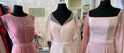Drei rosa Kleider auf Mannequins werden in einem Boutique ausgestellt, mit einer floralen Wand und einem Spiegel, der einen Raum mit einem Schrank und Blumen reflektiert.