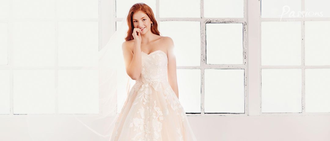 Eine Braut mit roten Haaren lächelt und posiert für ein Foto. Sie trägt ein weißes Hochzeitskleid mit langem Schleier. Der Hintergrund ist ein großes Fenster mit weißen Rahmen.