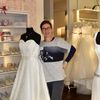 Eine Frau mit Brille und einer Uhr steht neben zwei Hochzeitskleidern auf Mannequins in einem Brautmodengeschäft. Im Hintergrund hängen weitere Hochzeitskleider an einem weißen Kleiderständer.