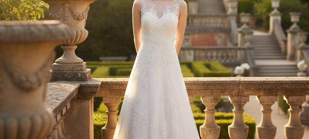 Eine Braut steht auf einem Balkon in einem Garten, trägt ein weißes Hochzeitskleid mit V-Ausschnitt und langer Schleppe. Dahinter führt eine Steintreppe zu einer Statue. Der Garten ist üppig grün.