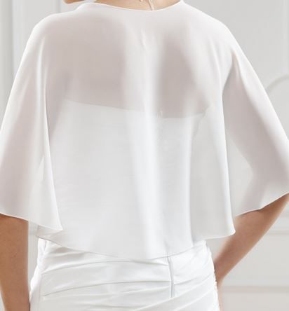 Eine Frau in einem weißen Kleid mit durchsichtigen Ärmeln steht gegen eine weiße Wand, ihre Hände liegen auf den Hüften.