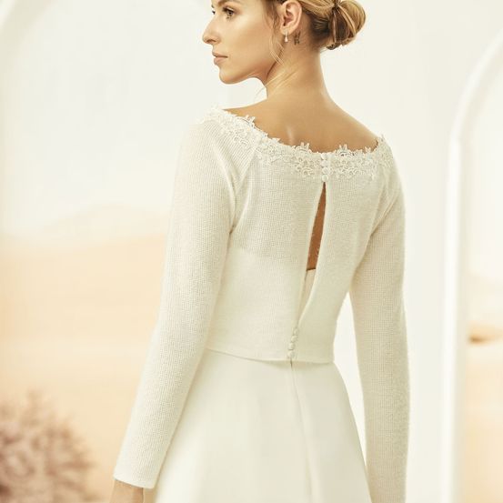 Eine Frau in einem Hochzeitskleid mit Spitzenschulterdetail steht vor einem weißen Hintergrund.