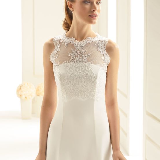 Eine Frau in einem weißen Hochzeitskleid mit aufwendigen Spitzendetails steht in einem hellen Raum.