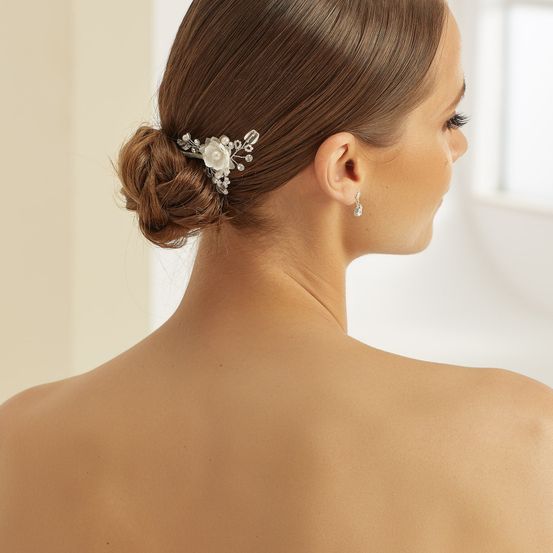 Eine Frau mit braunen Haaren, gestylt in einem Dutt, trägt eine silberne Blumenhaarnadel. Sie trägt Ohrringe und schaut nach rechts. Der Hintergrund ist unscharf.