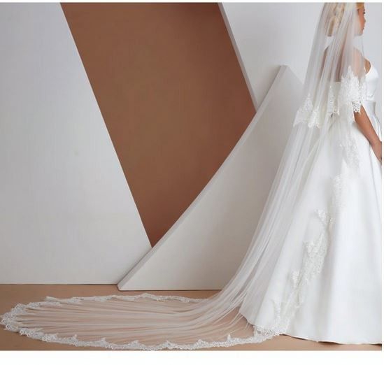 Eine Braut in einem weißen Kleid mit Spitzendetails und einem langen Schleier, steht in einem Raum mit weißen und braunen geometrischen Wänden.