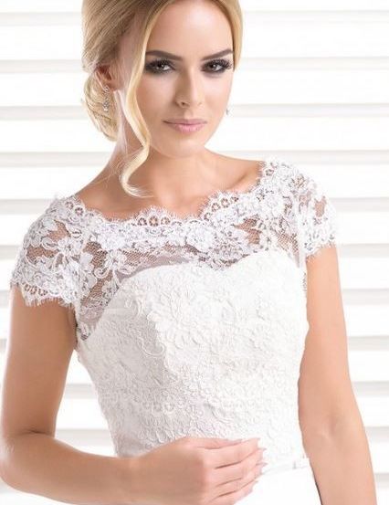 Eine Frau in einem weißen Hochzeitskleid mit Spitzenärmeln steht vor einem weißen Fenster. Sie hat blonde Haare und lächelt.