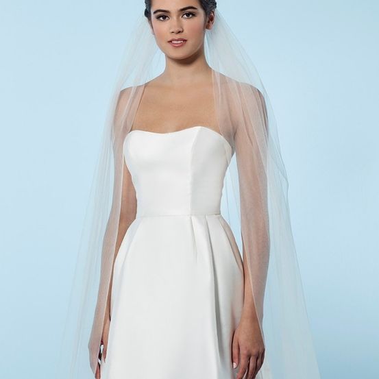 Eine Frau in einem weißen Hochzeitskleid mit langem Schleier steht vor einem blauen Hintergrund. Sie lächelt und scheint für ein Foto zu posieren.