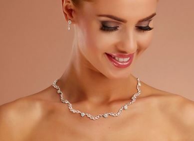 Eine Frau lächelt und trägt eine silberne Halskette mit Kristallen, ergänzt durch passende Ohrringe, vor einem pfirsichfarbenen Hintergrund.