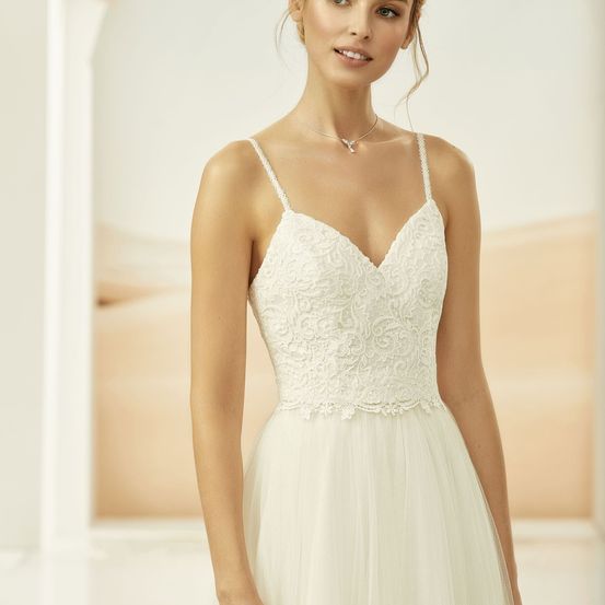 Eine Frau trägt ein weißes Spitzenhochzeitskleid mit V-Ausschnitt und fließendem Rock. Sie steht drinnen mit Wüstenhintergrund.