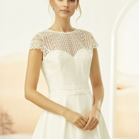 Eine Frau posiert in einem weißen Spitzenhochzeitskleid mit kurzen Ärmeln vor einem weich fokussierten Hintergrund.