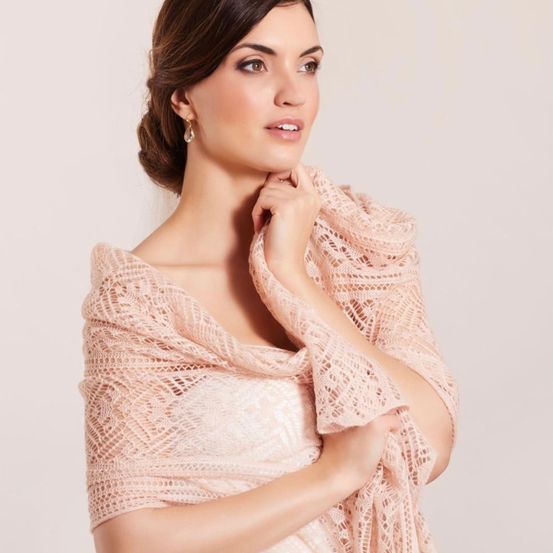 Eine Frau in einem weißen Kleid posiert für ein Foto, trägt einen hellrosa Schal mit komplizierten Spitzendetails. Sie hat braune Haare zu einem Dutt gebunden und trägt goldene Ohrringe.