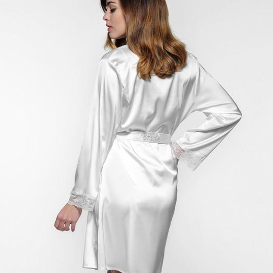 Eine Frau trägt einen weißen Seidenrob mit Gürtel und Spitzenbesatz, steht und schaut zur Seite.