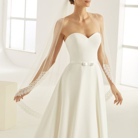 Eine Braut steht in einem weißen Raum und trägt ein weißes Kleid mit einem Spitzenschleier, ihre Hände halten elegant den Schleier.