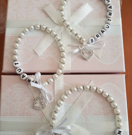 Zwei Perlenketten mit weißen Schleifen und Bögen liegen auf rosa Geschenkboxen mit Spitzendesigns. Die Halsketten haben silberne Anhänger und Perlen mit personalisierten Namen.