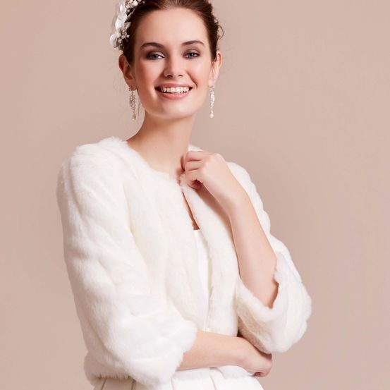 Eine lächelnde Frau mit einem weißen Pelzschal und Ohrringen posiert für ein Foto. Sie hat eine weiße Blume in den Haaren.