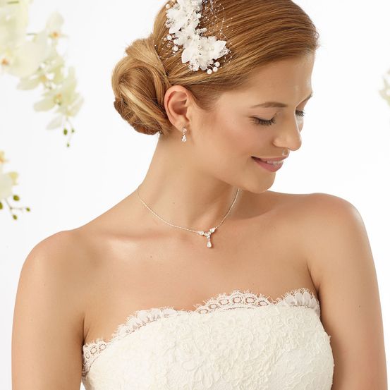 Eine Braut mit blondem Haar in einem Hochsteckfrisur, geschmückt mit einem weißen Blumenhaarzubehör und einer Perlenkette, blickt mit einem Lächeln nach vorne.