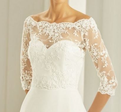 Eine Frau in einem weißen Hochzeitskleid mit Spitzenärmeln steht vor einem weißen Vorhang. Das Kleid hat einen herzförmigen Ausschnitt und aufwendige Spitzendetails.