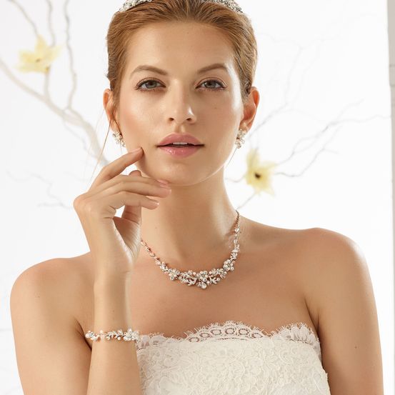 Eine Frau in einem weißen Hochzeitskleid und Perlenschmuck, die mit ernster Miene ihre Wange berührt, vor weißem Hintergrund.