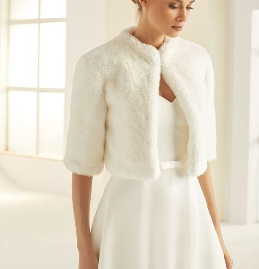 Eine Frau trägt ein weißes Kleid und einen Pelzmantel und steht vor einem Fenster.