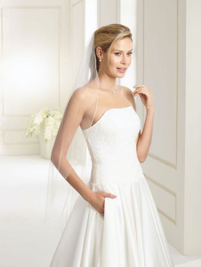 Eine Frau in einem weißen Hochzeitskleid und Schleier posiert für ein Foto in einem eleganten Raum mit weißen Wänden und einer Vase mit weißen Blumen.