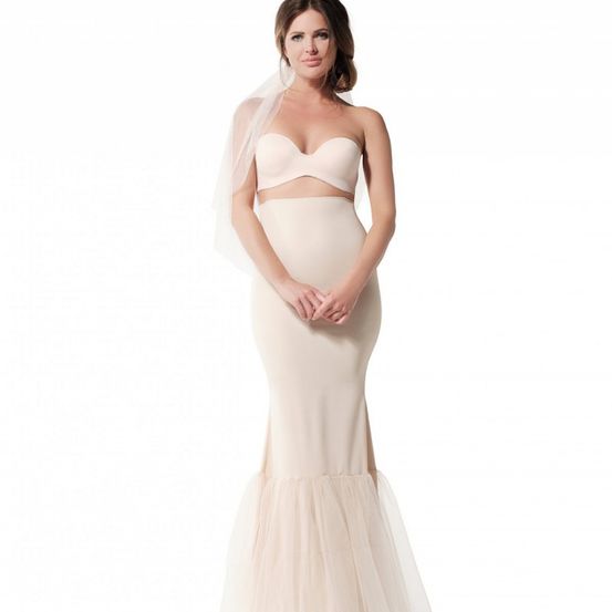 Eine Frau posiert in einem hellrosa Brautensemble, bestehend aus einem taillierten Oberteil und einem fließenden Rock mit Tüllüberlagerung. Sie trägt ein Schleier und hält ihre Hände anmutig.