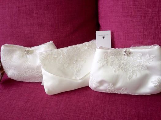 Drei weiße Spitzentaschen auf einer rosa Oberfläche. Jede Tasche hat dekorative Blumenspitze und eine kleine Kristallbrosche. Ein weißes Schild ist in der Mitte sichtbar.