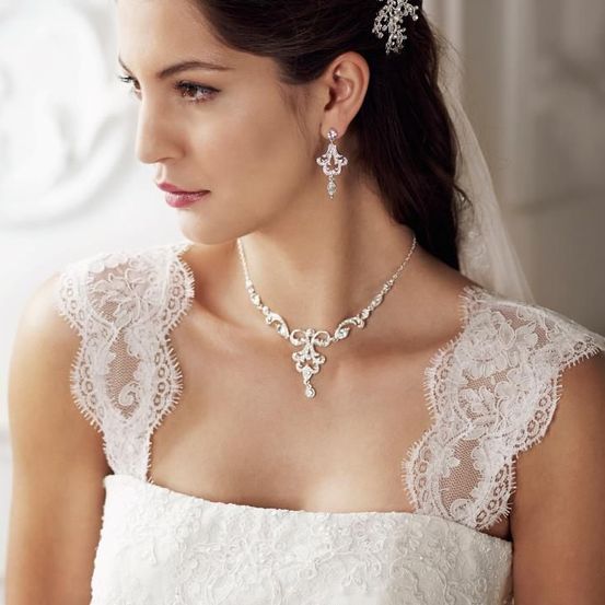 Eine Frau in einem Hochzeitskleid, Schleier und Schmuck posiert für ein Foto. Sie trägt ein Spitzenkleid mit aufwendigen Details, eine Halskette, Ohrringe und ein Haaraccessoire.