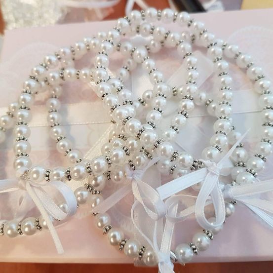 Drei Perlenarmbänder mit weißen Schleifen sind auf einer rosa Geschenkbox angeordnet. Die Armbänder haben silberne Akzente und weiße Perlen.